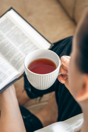 Close up, young woman drinking hot tea and reading favorite paper book.の写真素材