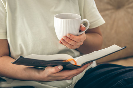 Woman holding a cup of tea and reading a book on the sofa at home, close up.の写真素材