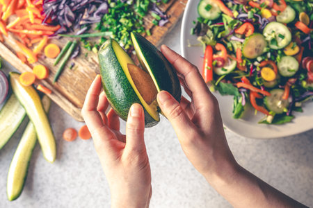 Avocado in female hands on a background of vegetables on the kitchen table.の写真素材