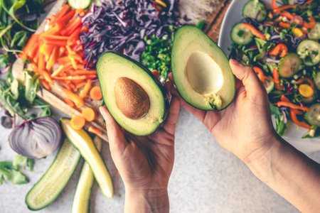 Avocado in female hands on a background of vegetables on the kitchen table.