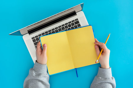 Open yellow notepad in female hands and a laptop on a blue background, top view.