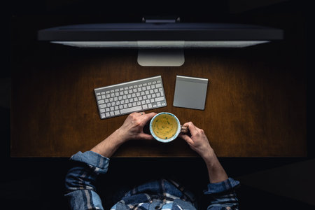 Top view of man working on computer at night, holding mug of tea.
