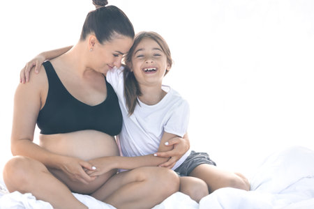 A pregnant woman and her teenager daughter are sitting on the bed.