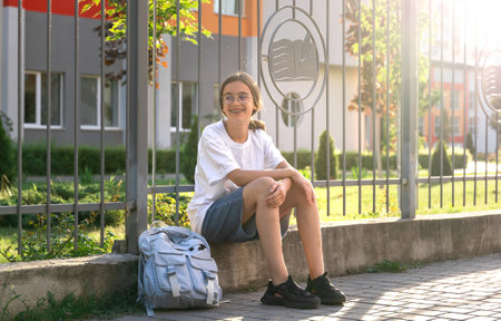 The student is sitting near the school fence. Schoolgirl with school supplies in hands. At sunset.の写真素材