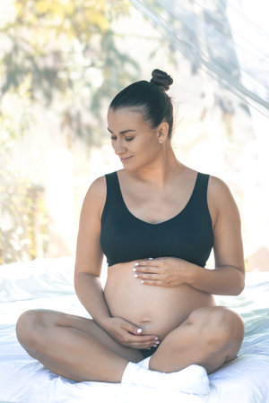 Happy pregnant woman touching her belly while sitting on a white bed.