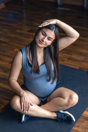 Pregnant woman in the gym doing warm-up. Sitting on a yoga mat.の写真素材