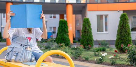 A girl with a blue notebook covers her face. On the background of the school.の写真素材