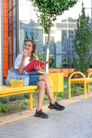 The student is sitting on a bench with a lunch box. Student with school supplies. Background school.の写真素材