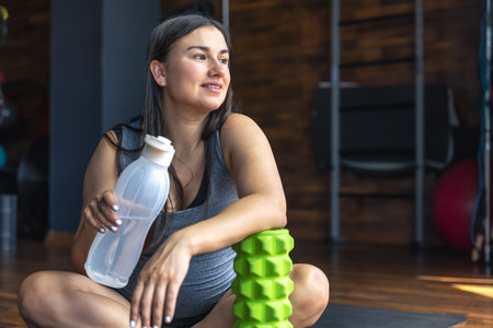 A pregnant woman in the gym sits on a mat and drinks water.の写真素材