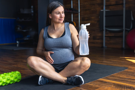 A pregnant woman in the gym sits on a mat and drinks water.の写真素材