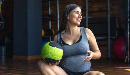 A pregnant woman in the gym sits on a mat with a ball in her hands.の写真素材