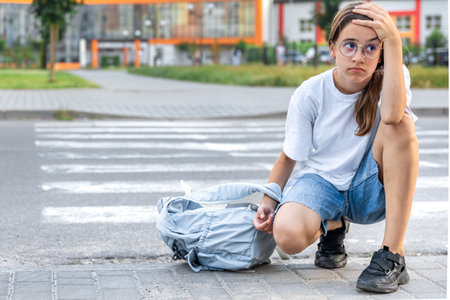 Upset schoolgirl by the road. Sitting at the crosswalk with a backpack. School background.の写真素材