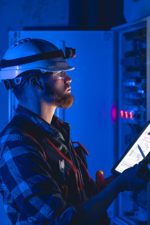 Electrician in overalls, focused on work in switchboard with fuses, using tablet.の写真素材