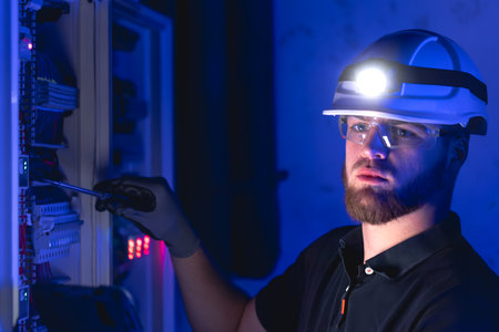 A male electrician works in a switchboard in overalls against the backdrop of emergency lighting.の写真素材