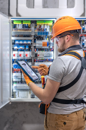 A male electrician in overalls, focused on work in switchboard with fuses, using tablet.の写真素材