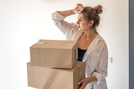 Young woman with boxes touches forehead in hallway. One person, moving.