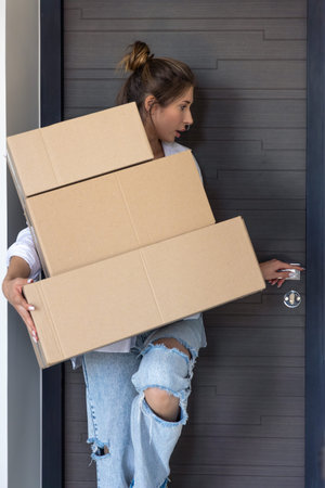 Young woman carrying three cardboard boxes at the front door. Moving and delivery.
