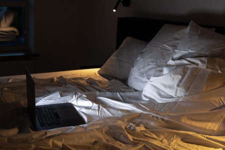 Side view laptop on rumpled bed under lamp. Late binge.