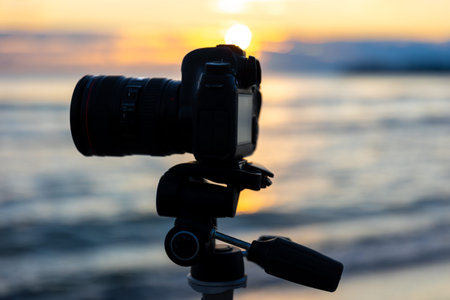 Close up of camera on tripod by sea waves at golden hour. Camera with lens.