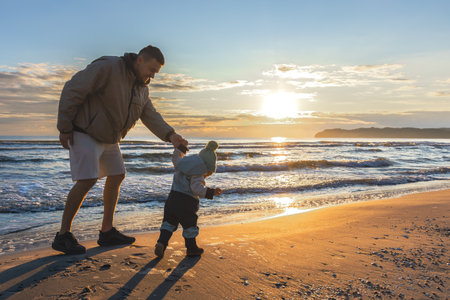 Dad walking toddler on beach in sunset light. Family joy by the sea.の写真素材