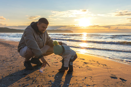 Toddler bends to sand while dad shows seashells. Family discoveries by the sea.の写真素材