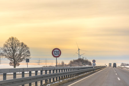 Winter highway with speed limit sign and wind turbines at sunset. Empty road landscape.