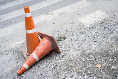 Two orange traffic cones on damage concrete road, one standing and the other one falling by...