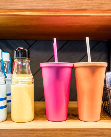 Easy grip glass water bottle standing by pink and orange stainless steel cold cup for cold beverages on wooden shelf against black tile background in coffee shop.の写真素材