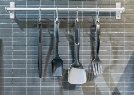 Essential kitchen utensils hanging with stainless hook on silver metal rail against black mosaic wall.の写真素材