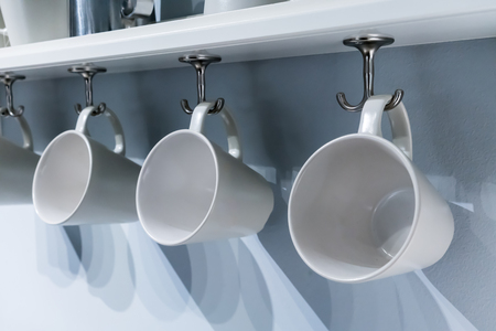 Row of white ceramic cups hanging on silver metal hook under white cabinet. Modern interior design.の写真素材
