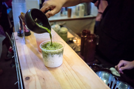 Woman hand holding black cup adding green milk tea on top of milk in clear plastic cup on wooden tableの写真素材