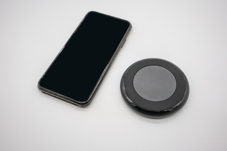Black mobile phone placed by black round shape wireless charger isolated on white background.