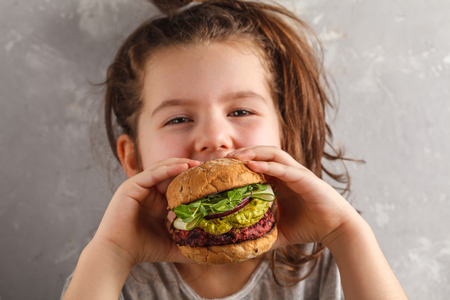 Beautiful happy hungry baby girl eating vegan burger.  Vegan beet chickpea burgers with vegetables, guacamole and rye buns. Healthy child vegan food concept.の写真素材
