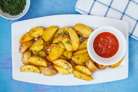 Baked potato slices with herbs in a white dish on a blue background, top  view. Vegan Healthy Food...