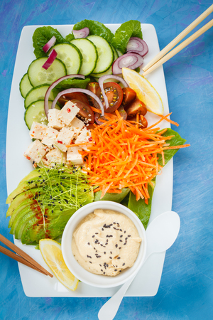 Rectangular rainbow vegan plate with vegetables, tofu and hummus. Vegetarian detox food concept. Blue background, top view.の写真素材