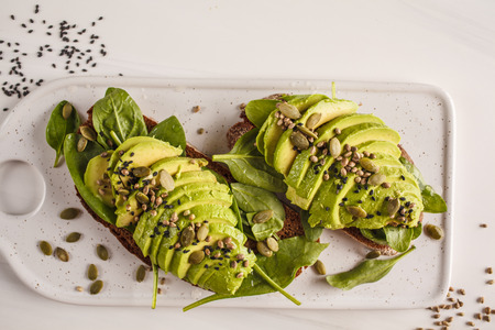 Avocado toast with hemp seeds, sesame seeds and pumpkin seeds on a white marble board, top view....