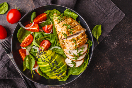 Grilled chicken breast and avocado salad with spinach, tomatoes and Caesar dressing in black plate, dark background.の写真素材