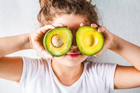 Healthy baby food concept. Girl child holds a sliced avocado in place of the eyes.の写真素材