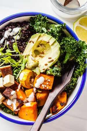 Buddha bowl salad with black rice, avocado, tofu, yam, kale and tahini dressing, white background.