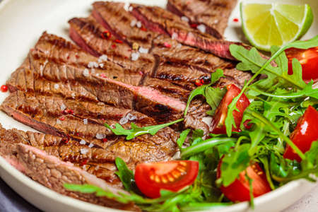 Sliced grilled beef steak with arugula and tomato salad in a white plate, dark background.の写真素材