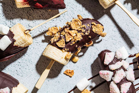 Bananas in dark chocolate with different toppings on a stick, gray background. Healthy vegan dessert concept.の写真素材