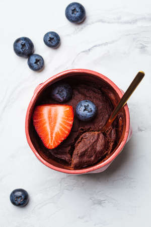 Chocolate mug cake with berries in a pink cup on a white marble background. Fast vegan microwave...