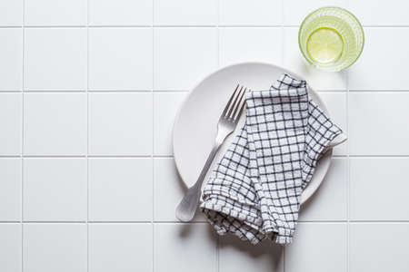Empty blank white plate with fork, napkin and glass of water on white tile background, copy space, top view. Interval fasting, healthy lifestyle, diet concept.の写真素材