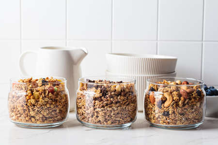 Different types of homemade granola (fruit, nut and chocolate) in jars on a white kitchen table. Breakfast food concept.の写真素材