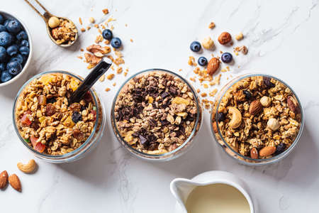 Different types of homemade granola (fruit, nut and chocolate) in jars on a white kitchen table, top...