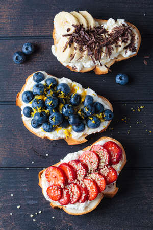 Fruity vegetarian toasts for breakfast on dark wooden background, top view. Bread slices with ricotta, berries, banana, chocolate and seeds.の写真素材