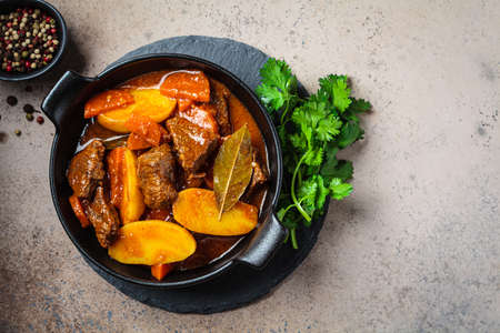 Portion of beef stew with potatoes, carrots in tomato sauce in black pot, dark background, top view.