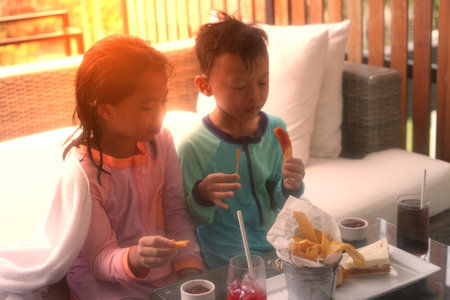 Boy and girl eat snack while rest time after play water on poolside sofa in summer vacation concept with sun lightの写真素材