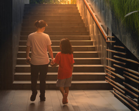 Mom and young girl hold hands walking along way to stair head to beach front in summer vacation time for happiness travel tour family concept with sun lightの写真素材