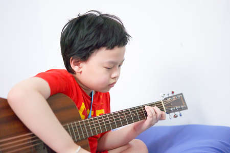 The boy plays the guitar with his left hand, a song by himself, on the sofa, in the bedroom, reading the sheet music.の写真素材